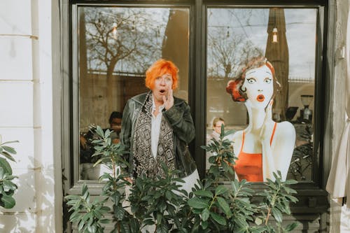 A woman with orange hair and a mannequin in front of a window