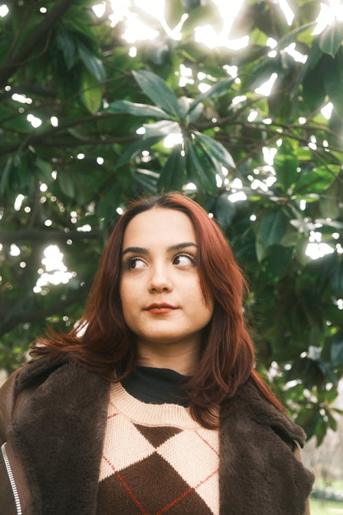 A woman with red hair and brown sweater is standing in front of trees