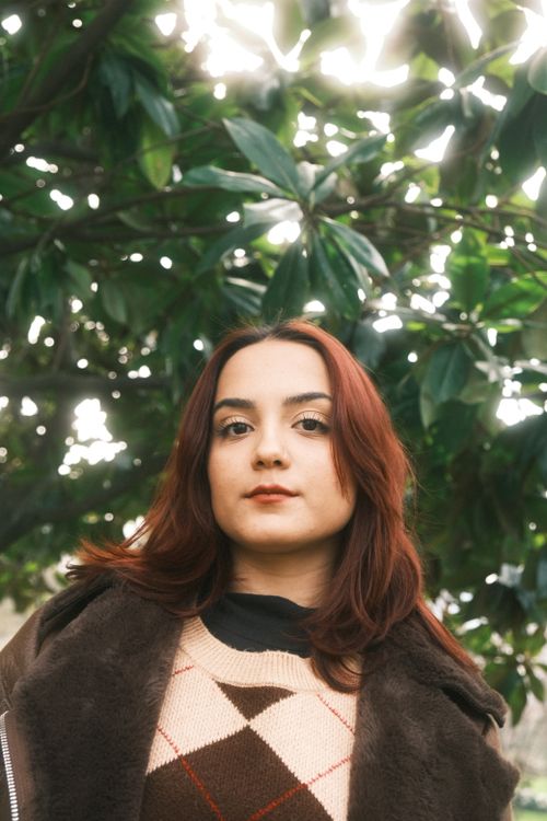 Free Portrait of a fashionable woman in an argyle sweater with lush green leaves in the background, showcasing a natural look. Stock Photo