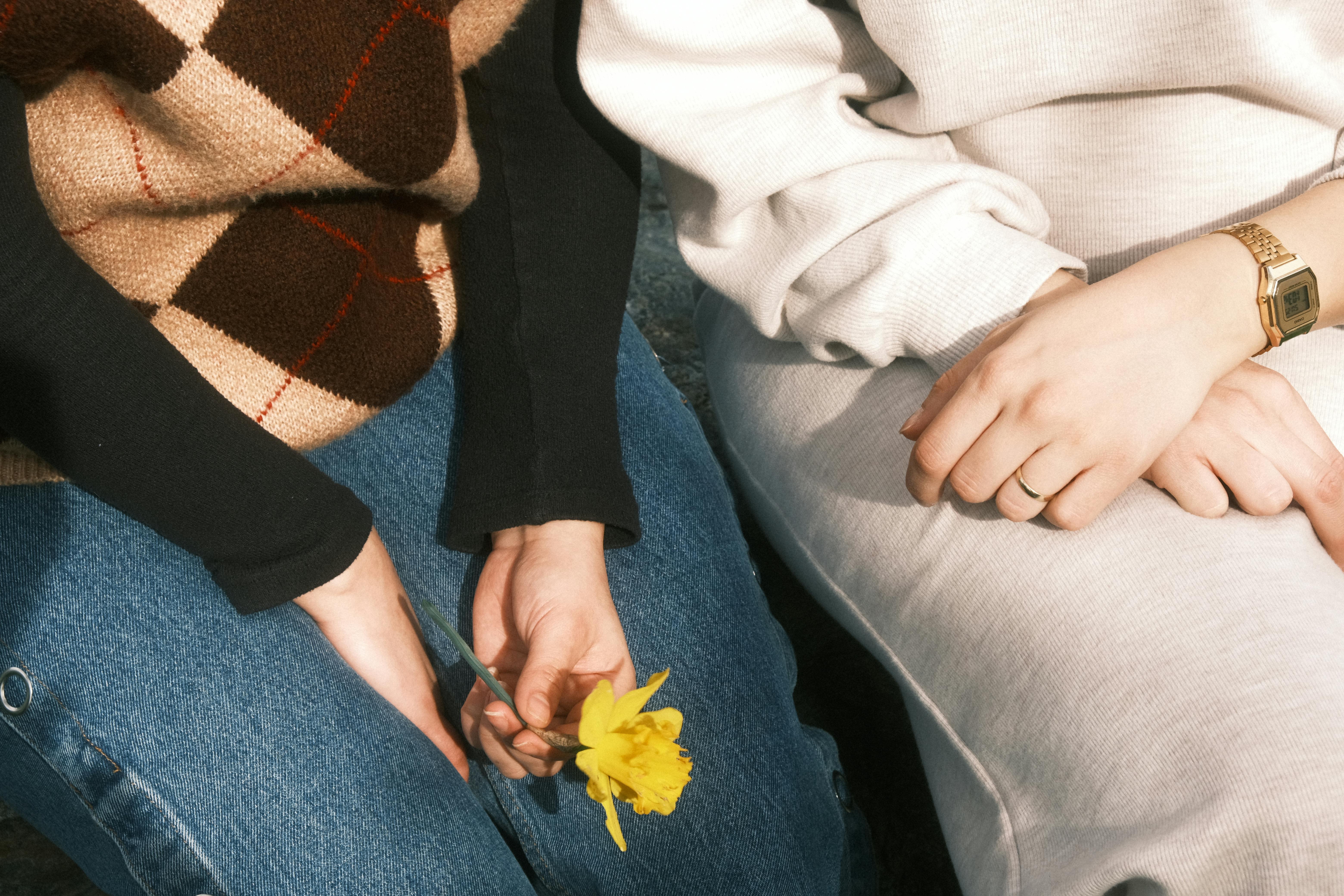 Two women sit side by side, one holding a yellow flower, showcasing casual fashion.
