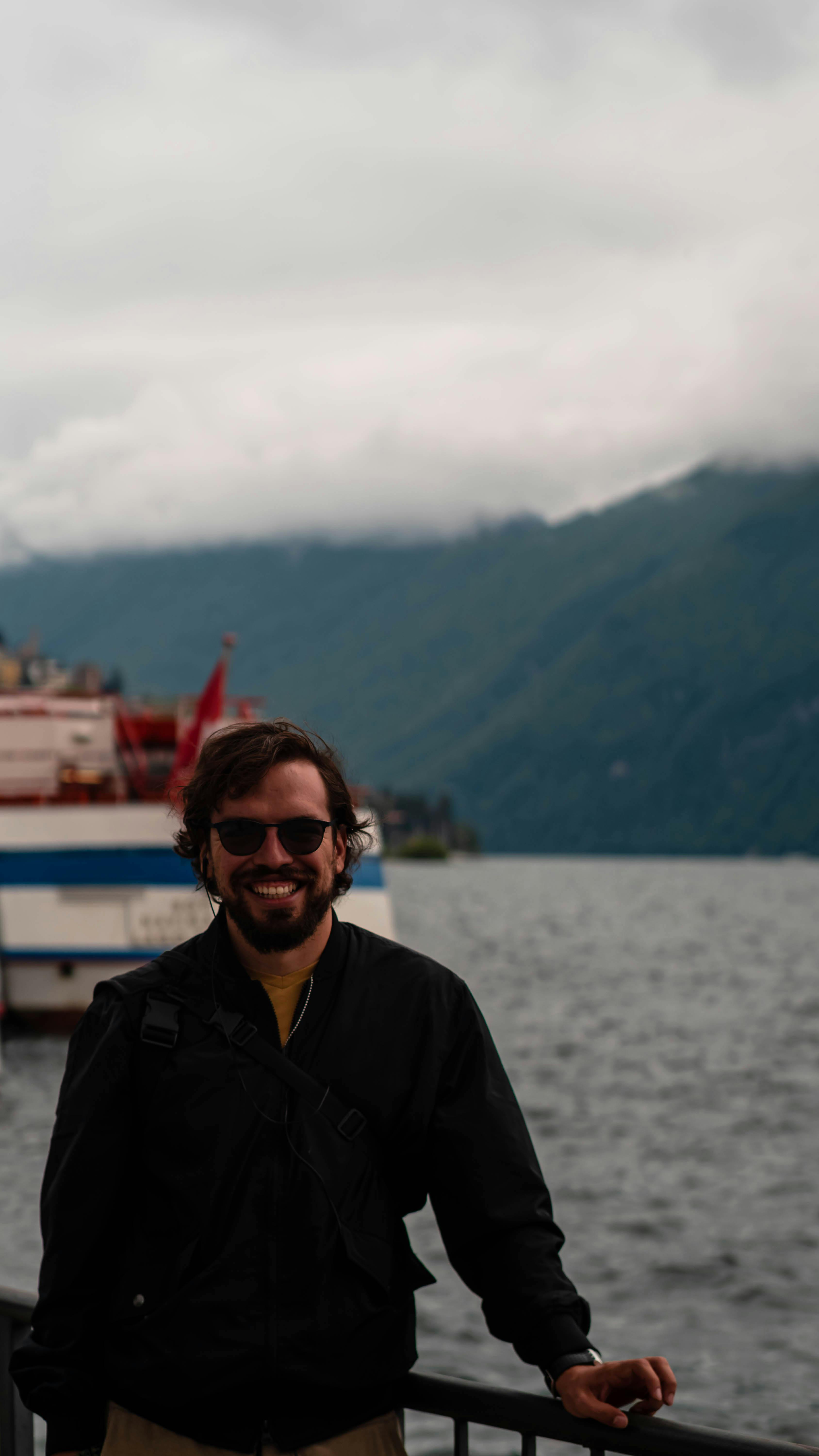 A smiling man in sunglasses stands by Lake Lugano with a ship in the background.