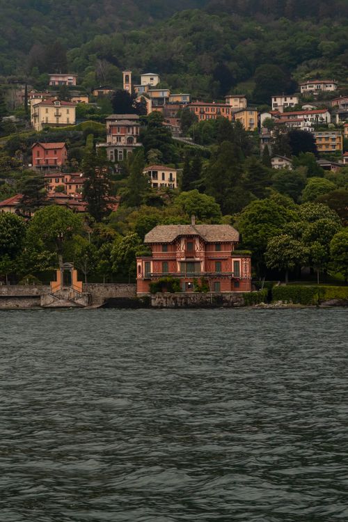 Free Picturesque lakeside view of a red house on Lake Como, Italy, surrounded by lush hills. Stock Photo