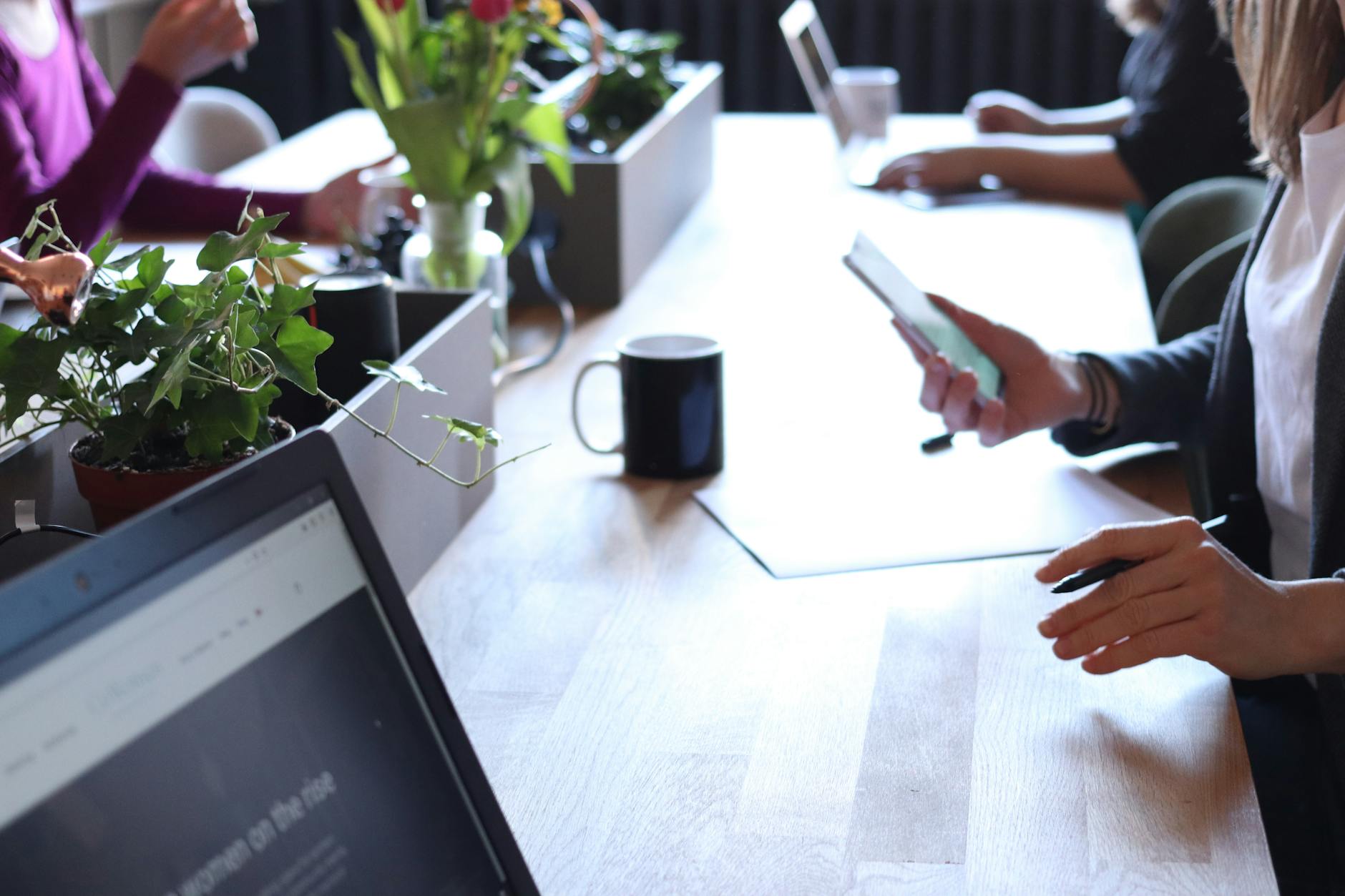 Office team collaborates around table; laptops open, hand grips phone, fresh plants nearby.