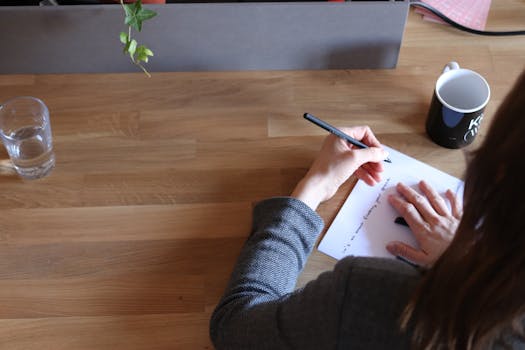 一名成年妇女在办公室的木制桌子上做笔记，桌上放着一个杯子和一杯水