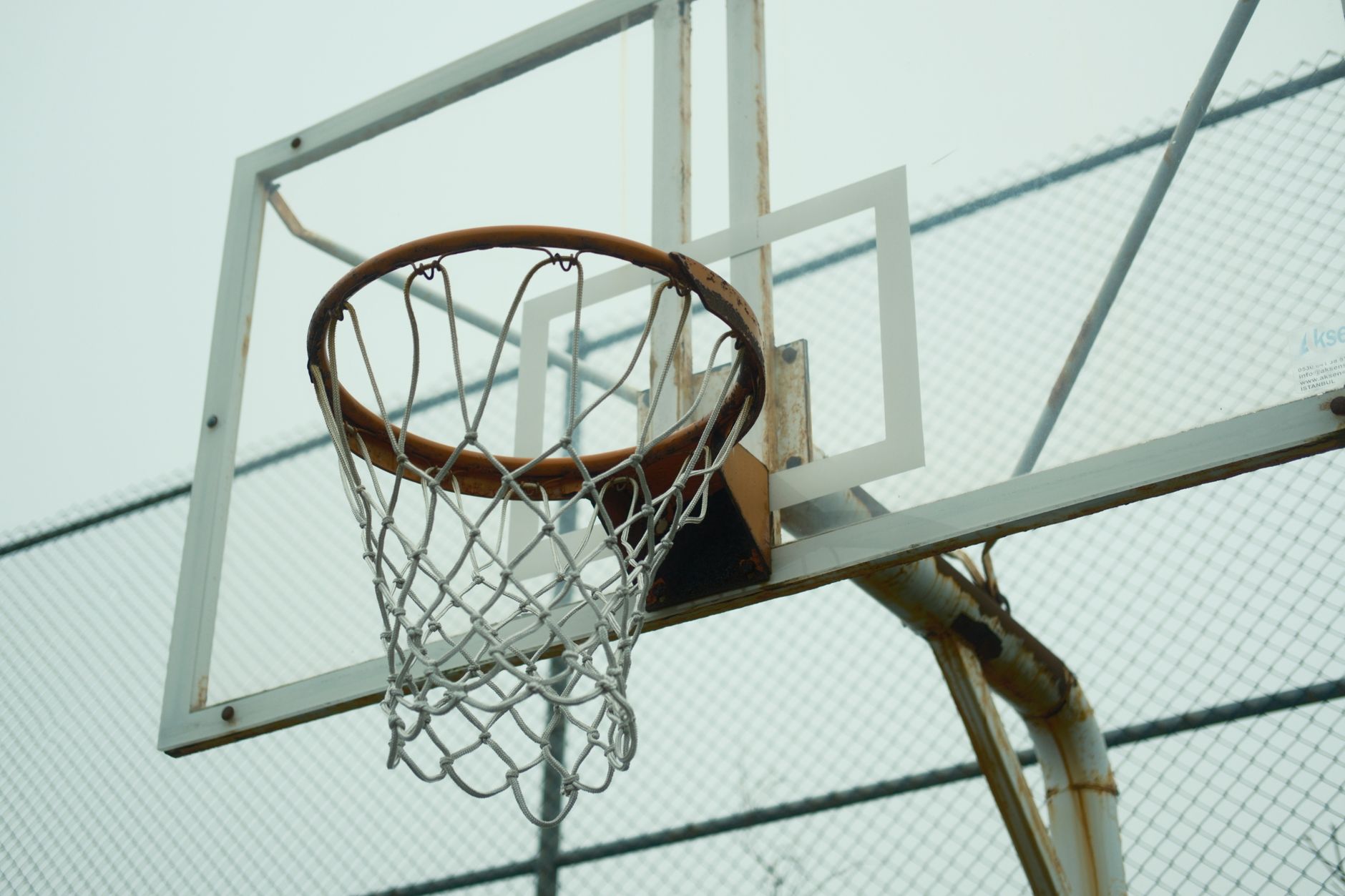 Close-up of an outdoor basketball hoop against a chain-link fence and overcast sky.