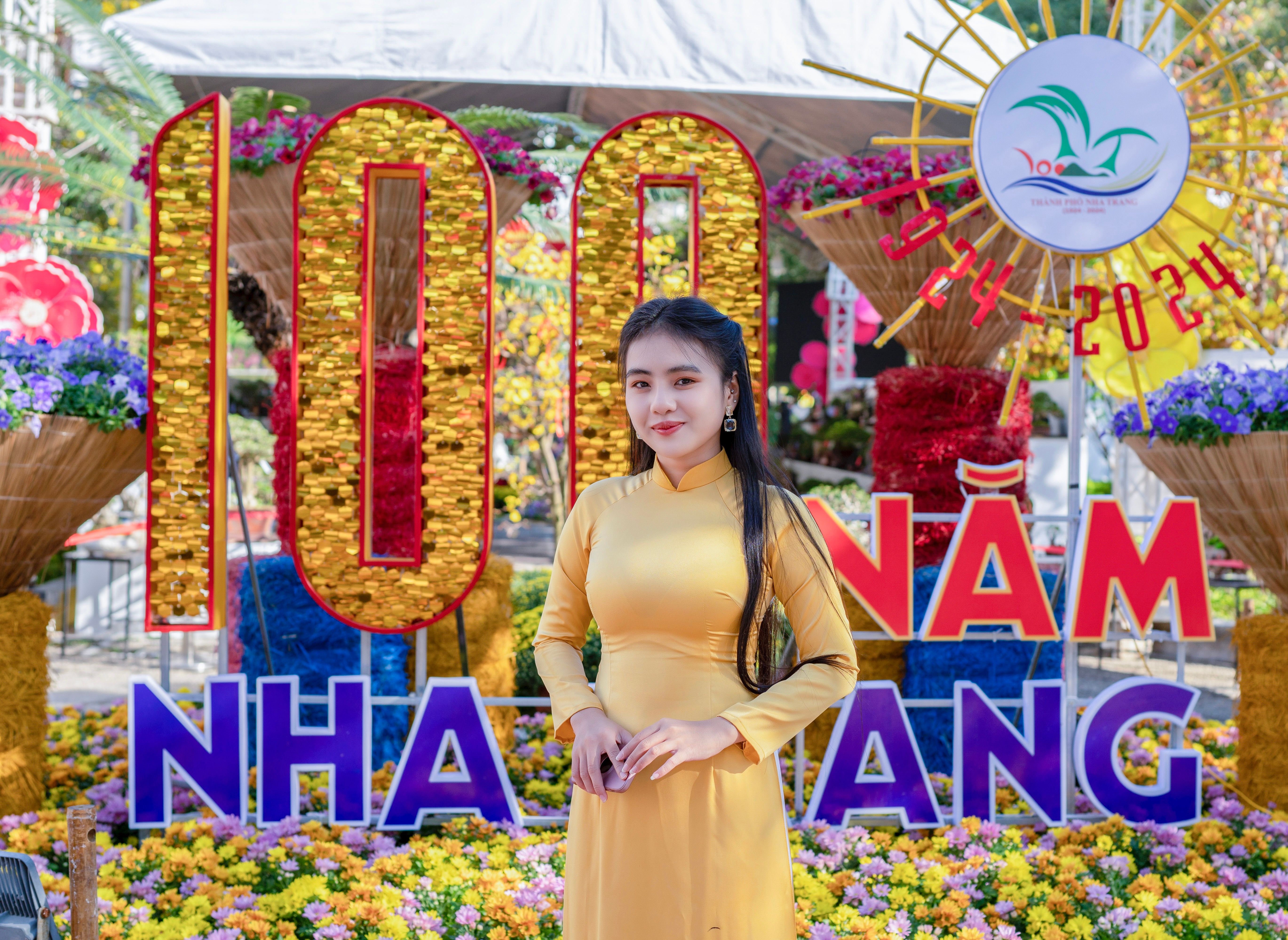 A Vietnamese woman in a yellow dress posing by a festive floral display in an urban setting.