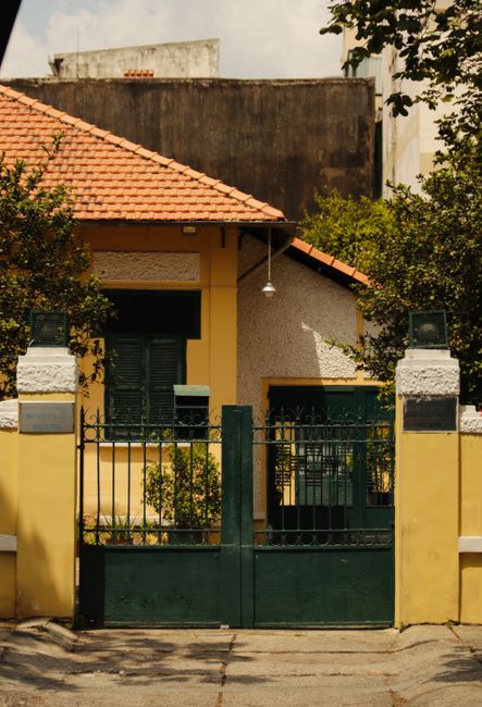 A quaint yellow house with a red-tiled roof and green gate surrounded by lush trees.