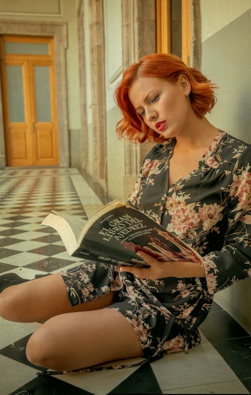 A woman with red hair is sitting on the floor reading a book