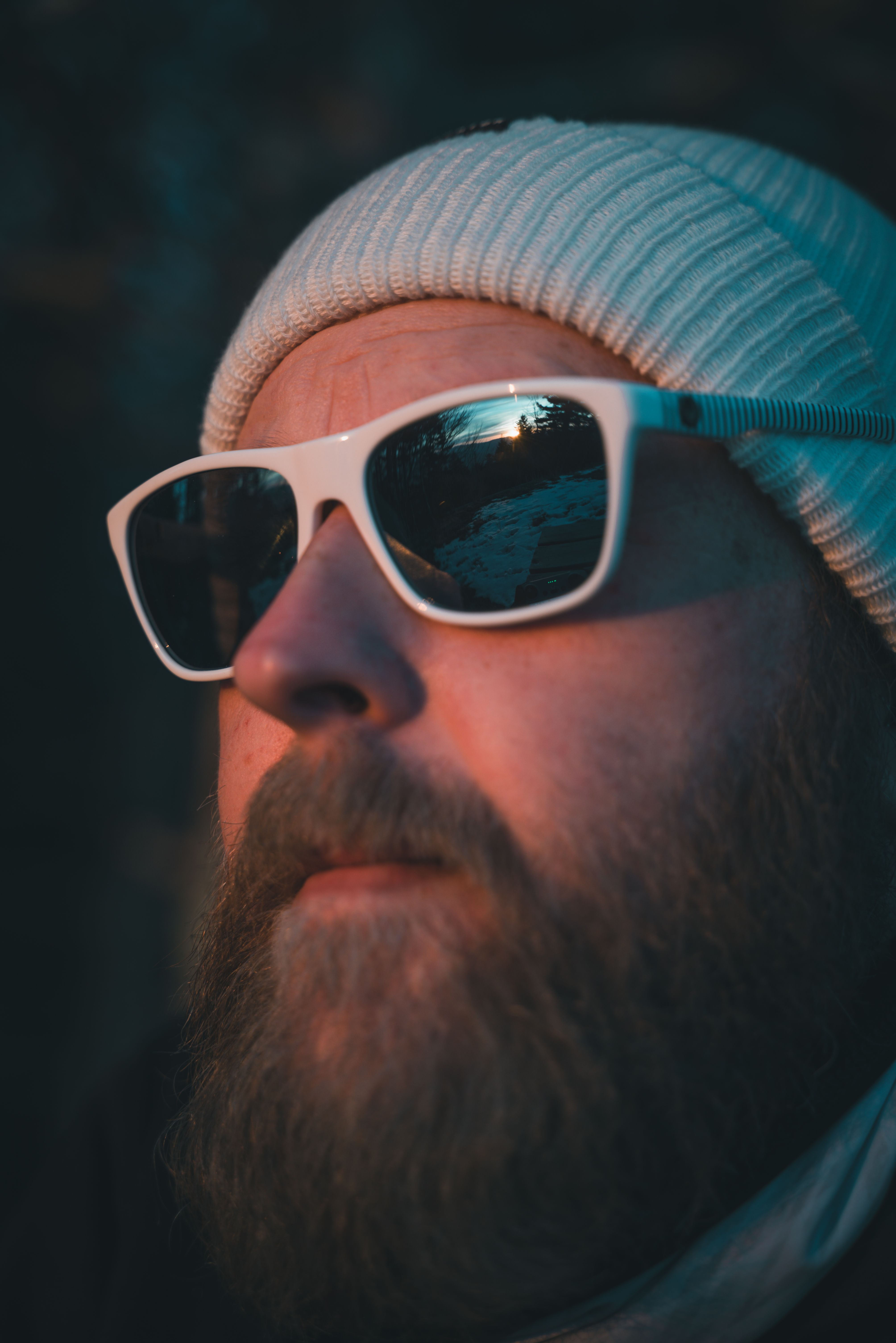 Portrait of a bearded man wearing white sunglasses and a beanie, captured in warm evening light.