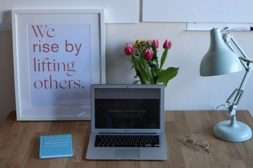 Free Comfortable desk with opened netbook book and cozy lamp placed near framed image with inscription we raise by lifting others Stock Photo