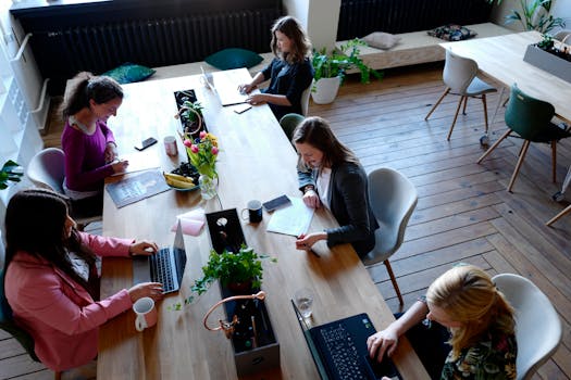 A high-angle shot of women working collaboratively in a modern office environment with laptops and plants.