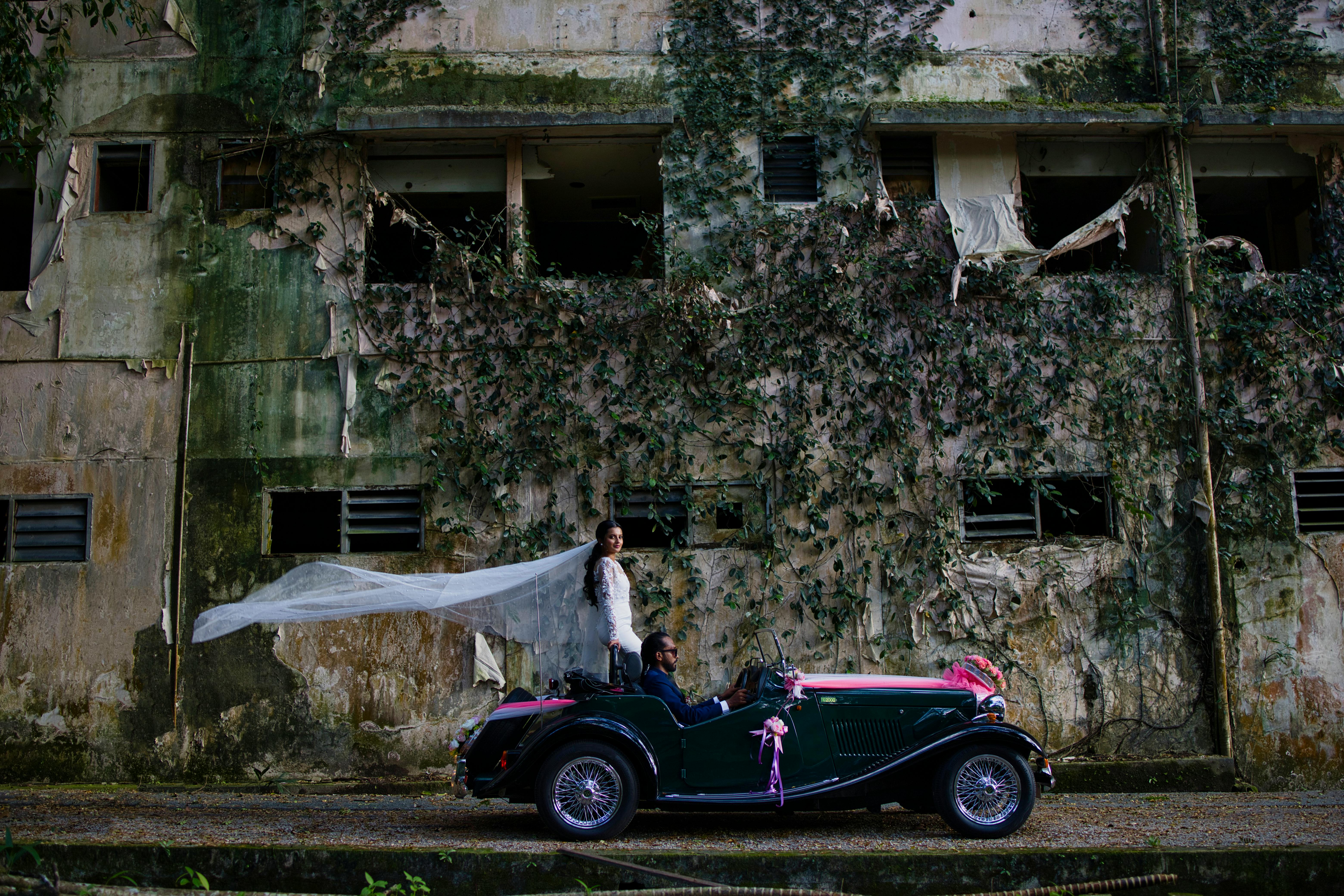 Bride and groom in classic car pose against rustic urban facade in Johor Bahru, Malaysia.