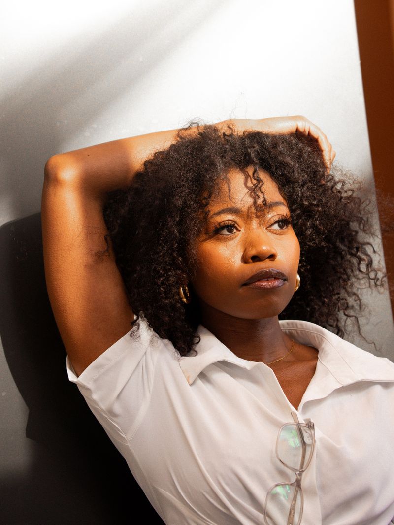 A Black woman with curly hair photographed in soft natural light indoors, a contemplative portrait used to illustrate reporting on lupus symptoms and diagnosis in Black women.