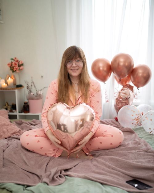 Free Woman in cozy pink pajamas holding a heart balloon sitting on bed surrounded by balloons and flowers. Stock Photo