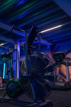 A modern gym filled with exercise equipment under blue lighting, featuring a stationary fan bike.