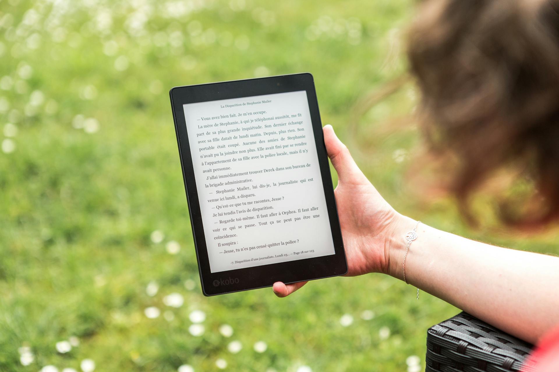 A person reads an eBook on a Kobo device while relaxing outdoors in a sunny park.