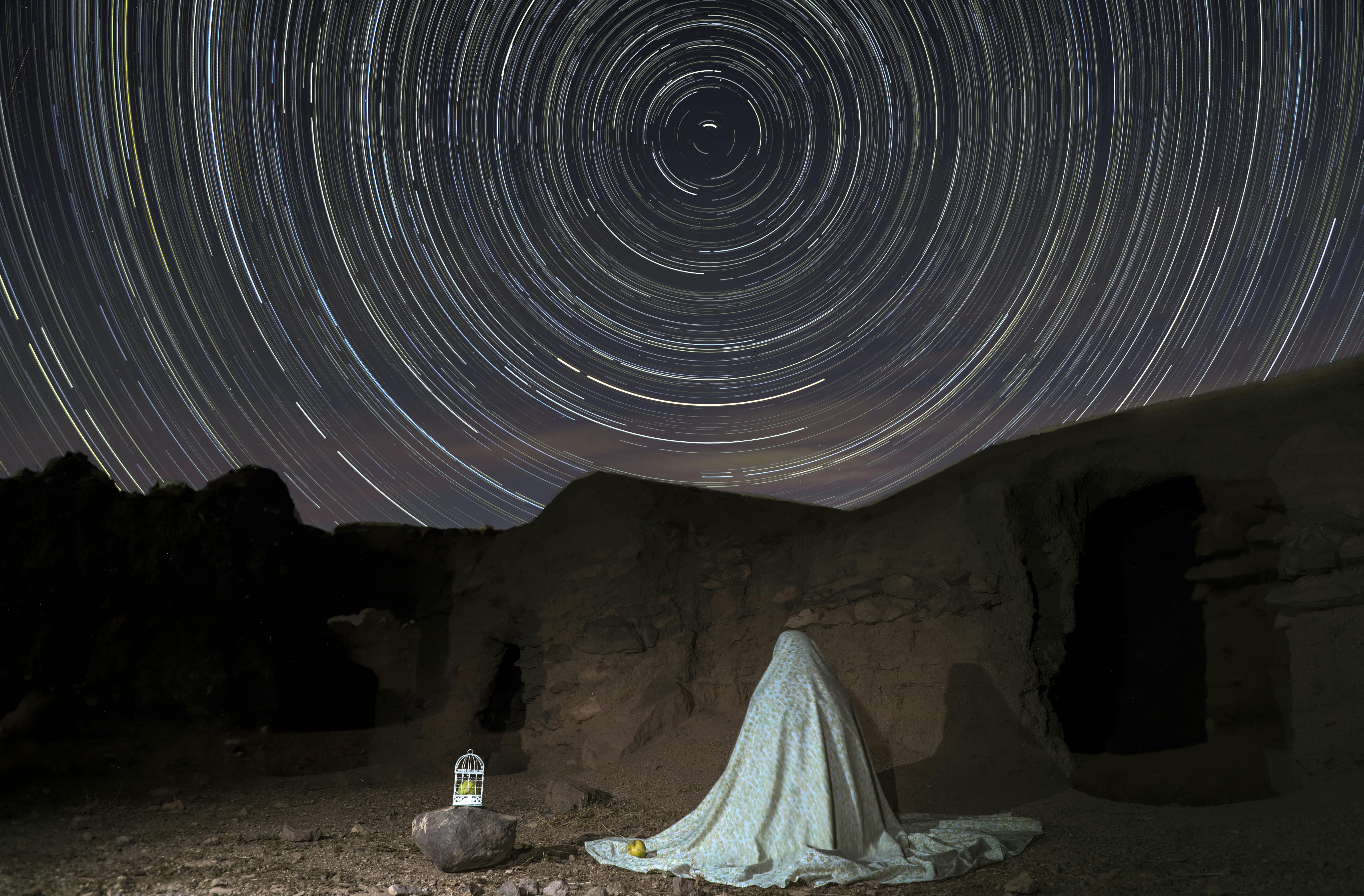 A person sits under a starry sky with vivid star trails, creating a mystical and serene atmosphere.