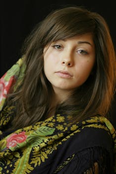 A striking portrait of a young woman with brown hair in a floral scarf, set against a dark background.