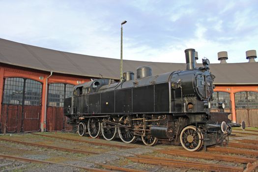 A beautifully preserved vintage steam locomotive stands in front of a historic roundhouse.