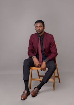 Black male model in a red suit poses confidently on a wooden chair in a professional studio setting.
