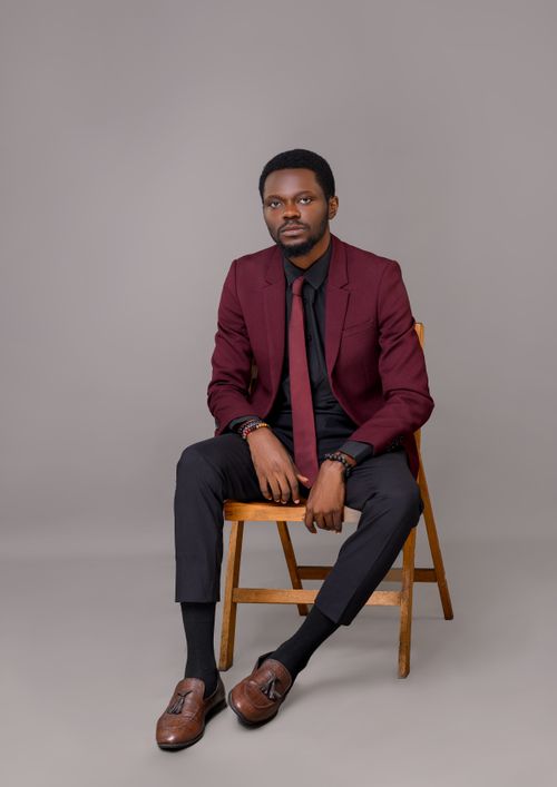Free Black male model in a red suit poses confidently on a wooden chair in a professional studio setting. Stock Photo