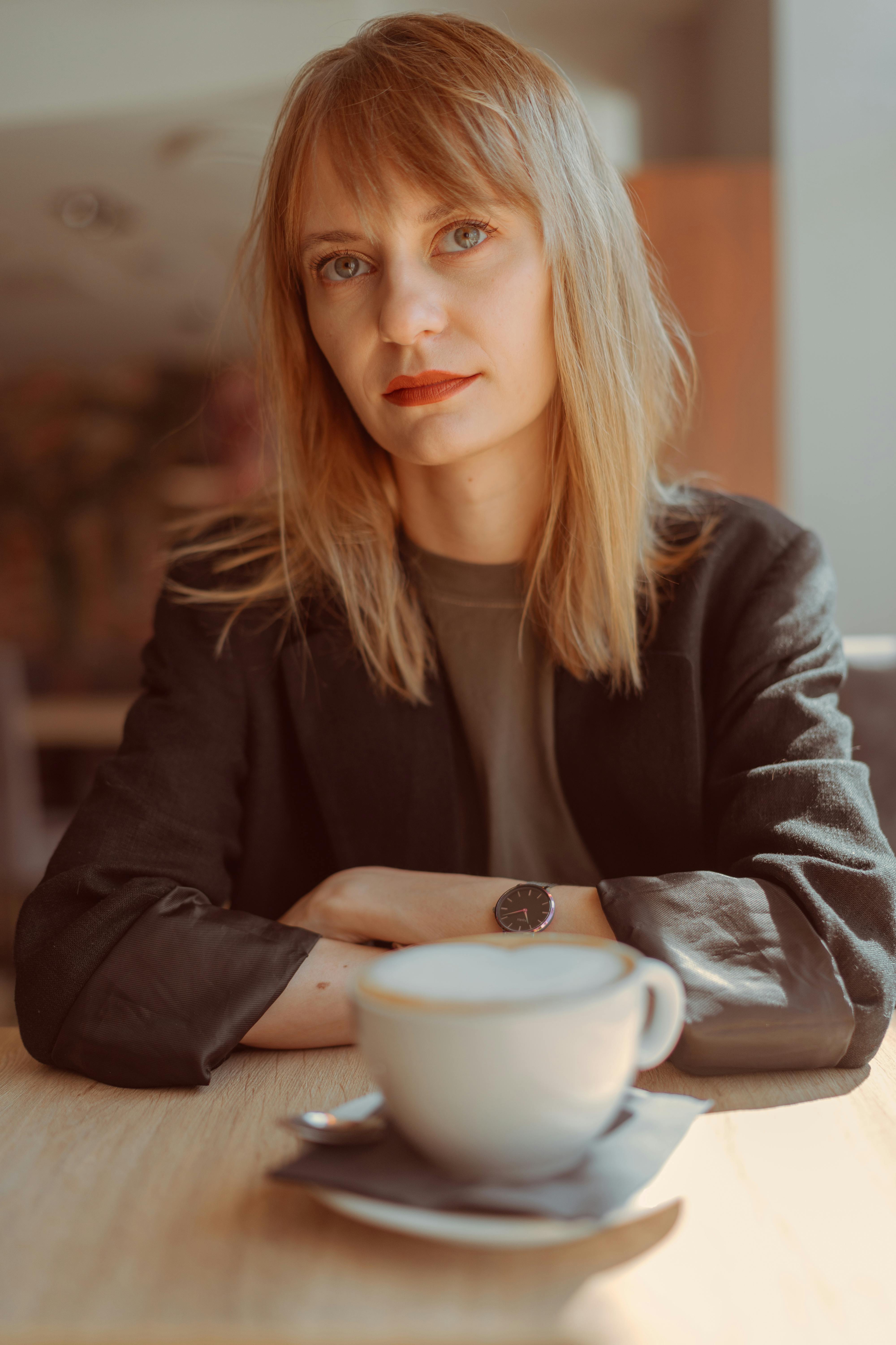 Blonde woman with long hair sits by a coffee cup in a cozy cafe setting.