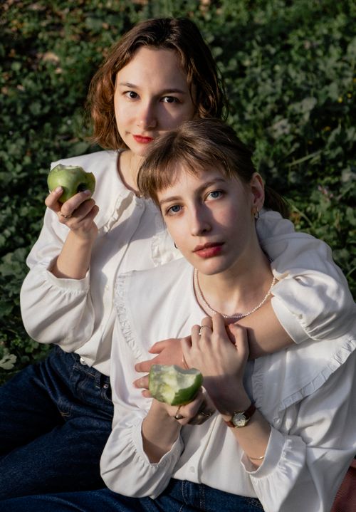 Free Two young women relaxing outdoors in white blouses, enjoying green apples on a sunny day. Stock Photo