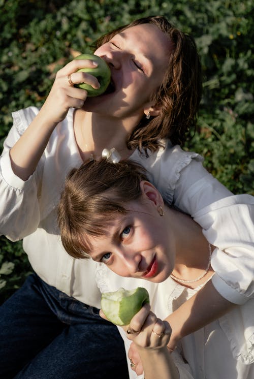 Two women are sitting on the grass eating apples