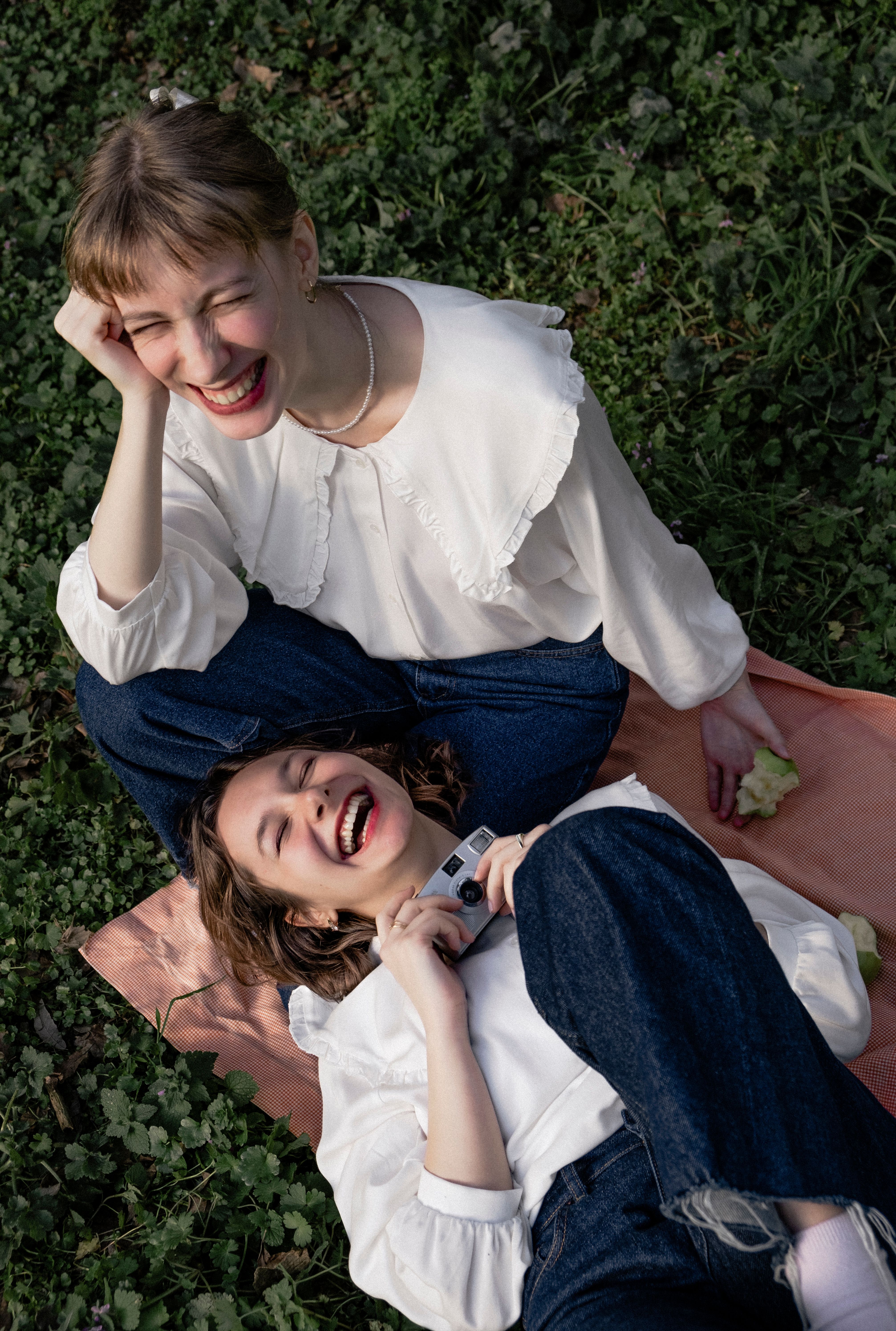 Two women joyfully laughing while lying on a picnic blanket in casual fashion outdoors.