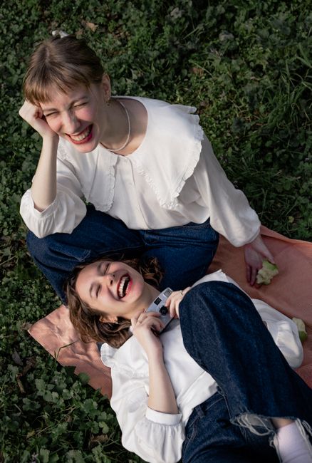 Two women joyfully laughing while lying on a picnic blanket in casual fashion outdoors.