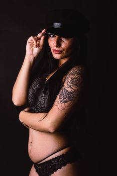 Studio portrait of a woman in black lingerie and hat, showcasing intricate tattoos and confident pose.