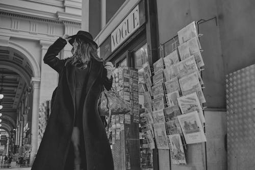 A stylish woman in a black coat and hat poses outdoors in a city street scene.