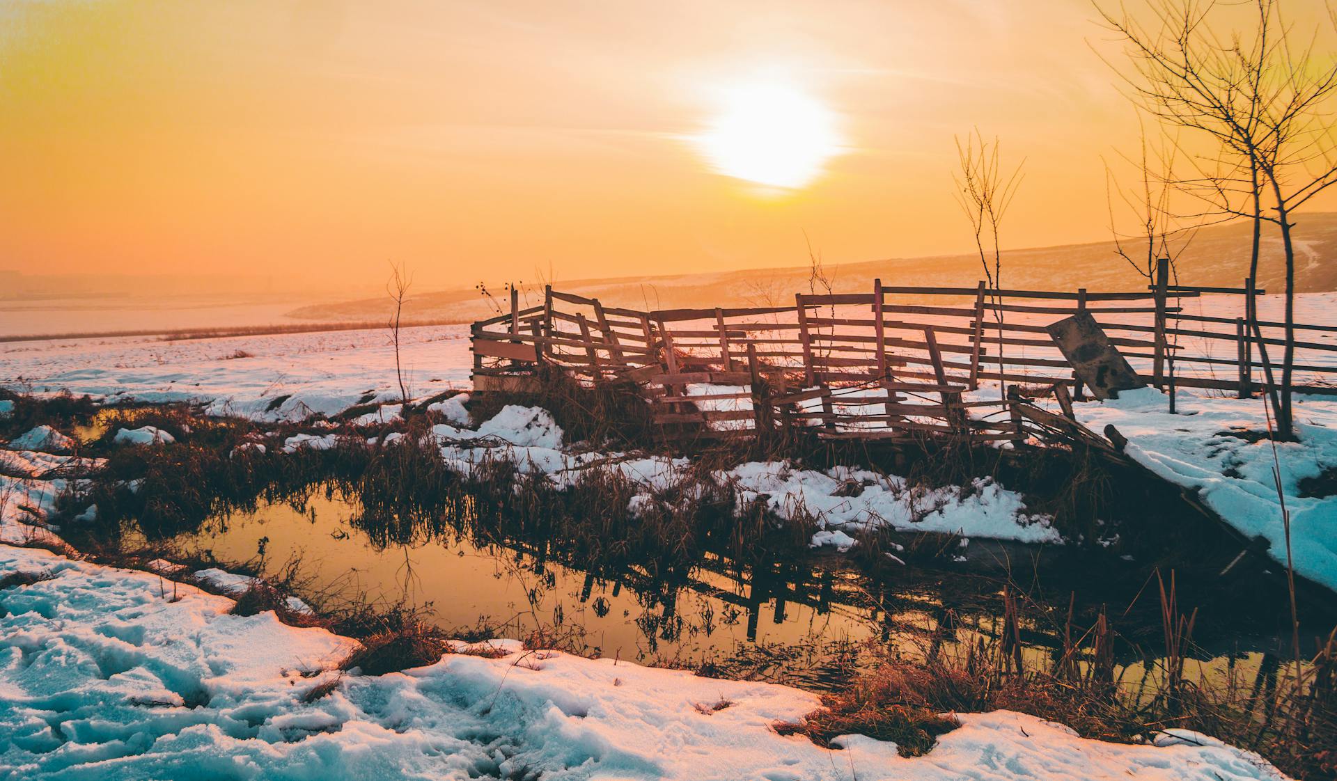 Idyllic winter scene with a wooden fence, snow, and setting sun near a stream in Kosovo Polje.