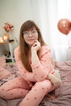 Woman in pink heart pajamas sitting in a cozy bedroom with balloons and decor.