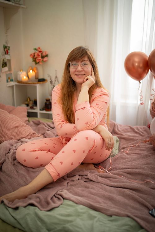 Free A smiling woman in pink pajamas sitting comfortably on a bed in a warmly lit bedroom. Stock Photo