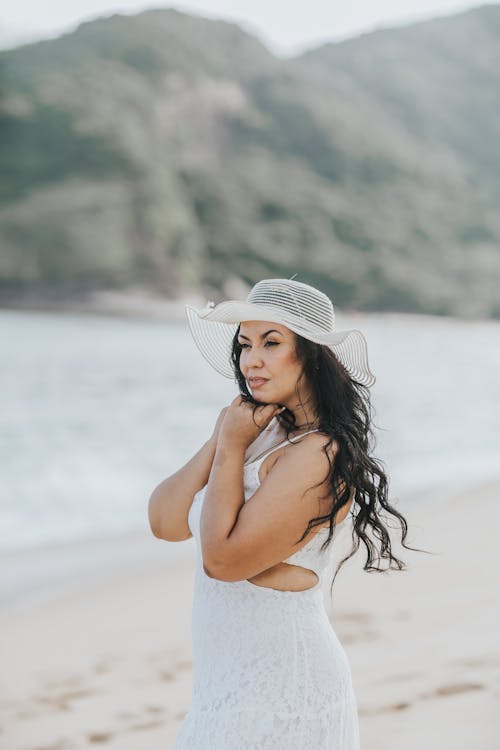 Woman in a White Dress and White Hat Standing on a Beach 