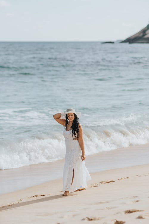 Woman in a White Dress and White Hat Walking on a Beach 