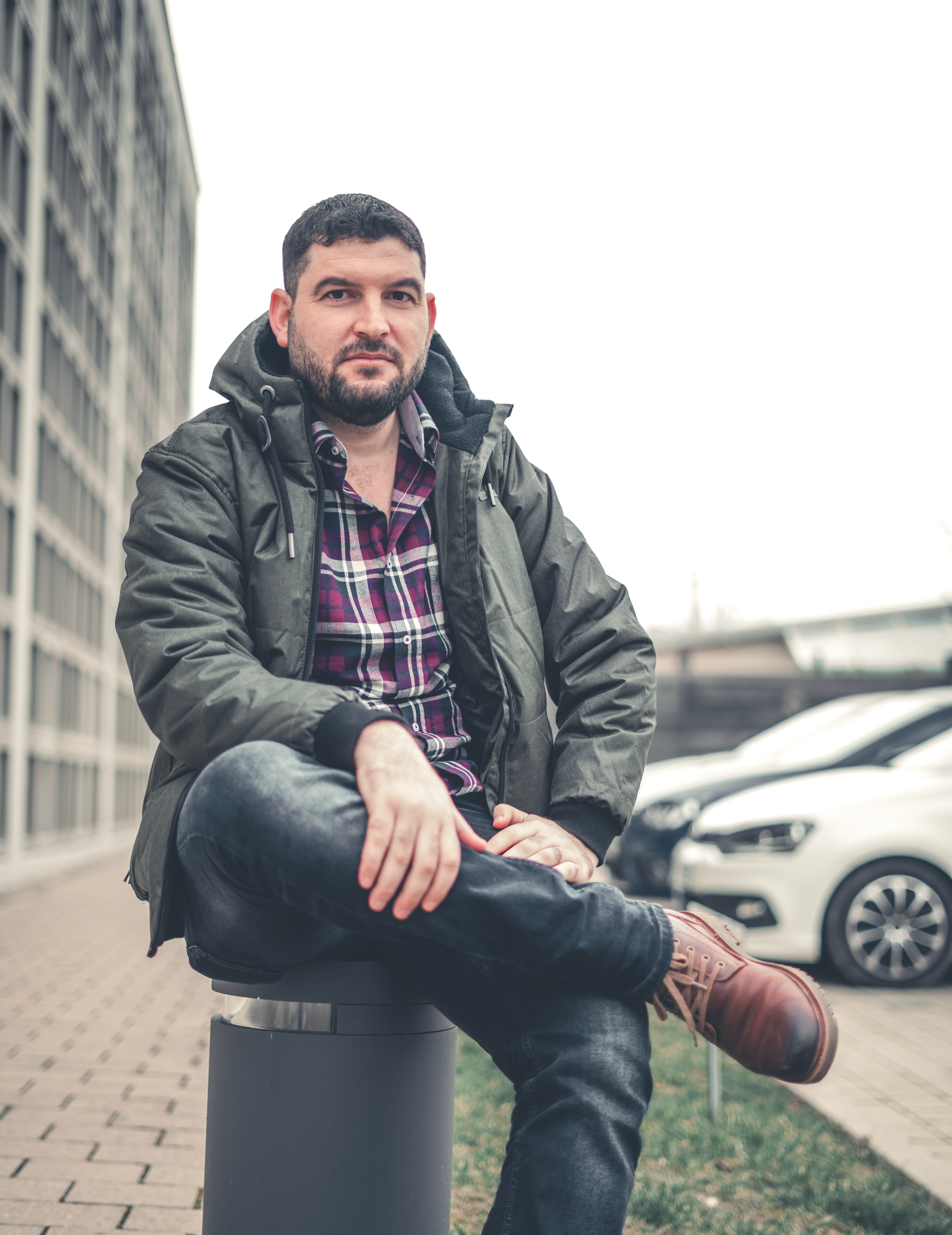 Casual man in green jacket and plaid shirt sitting outdoors in urban setting.