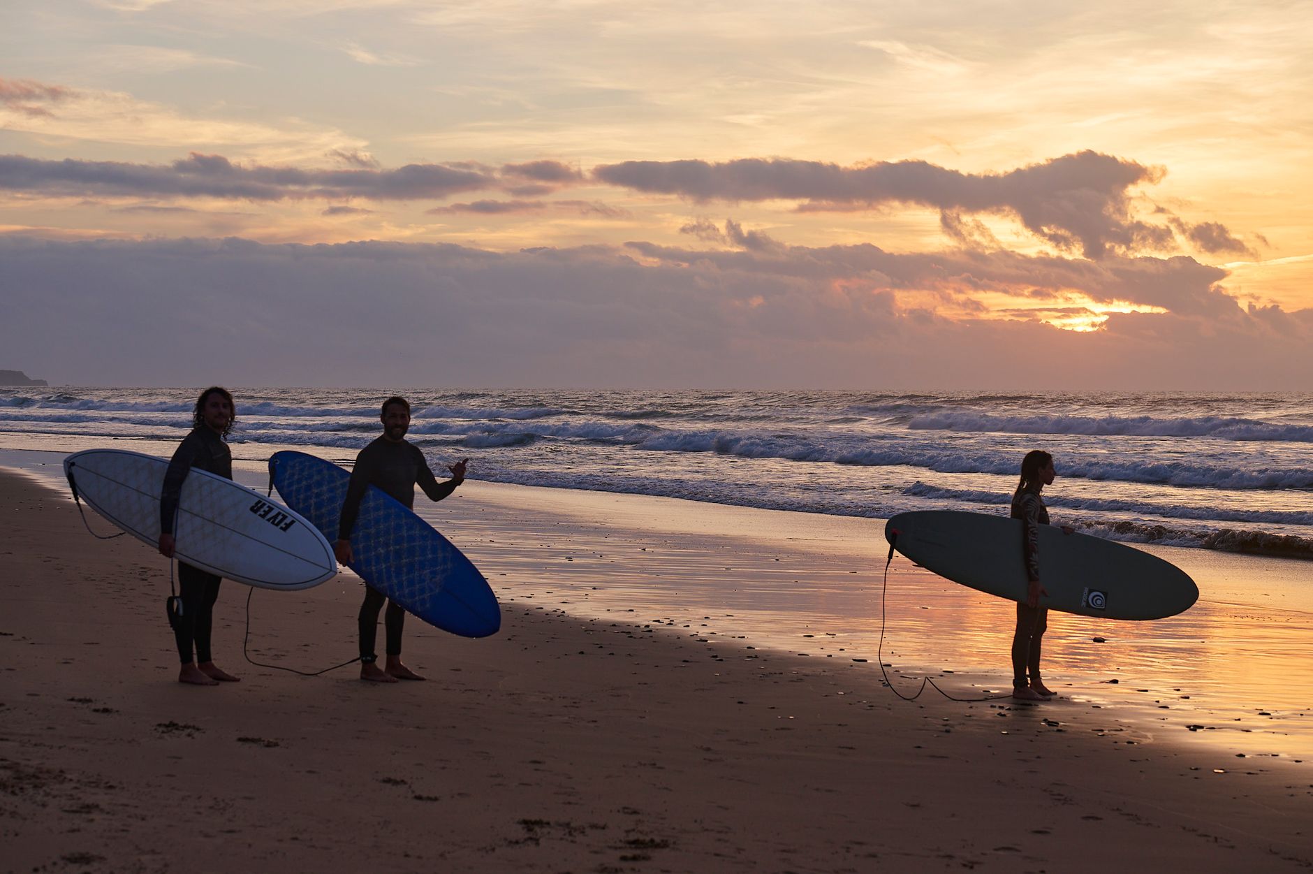 Three surfers with boards standing on a beach at sunset, ready to hit the waves.