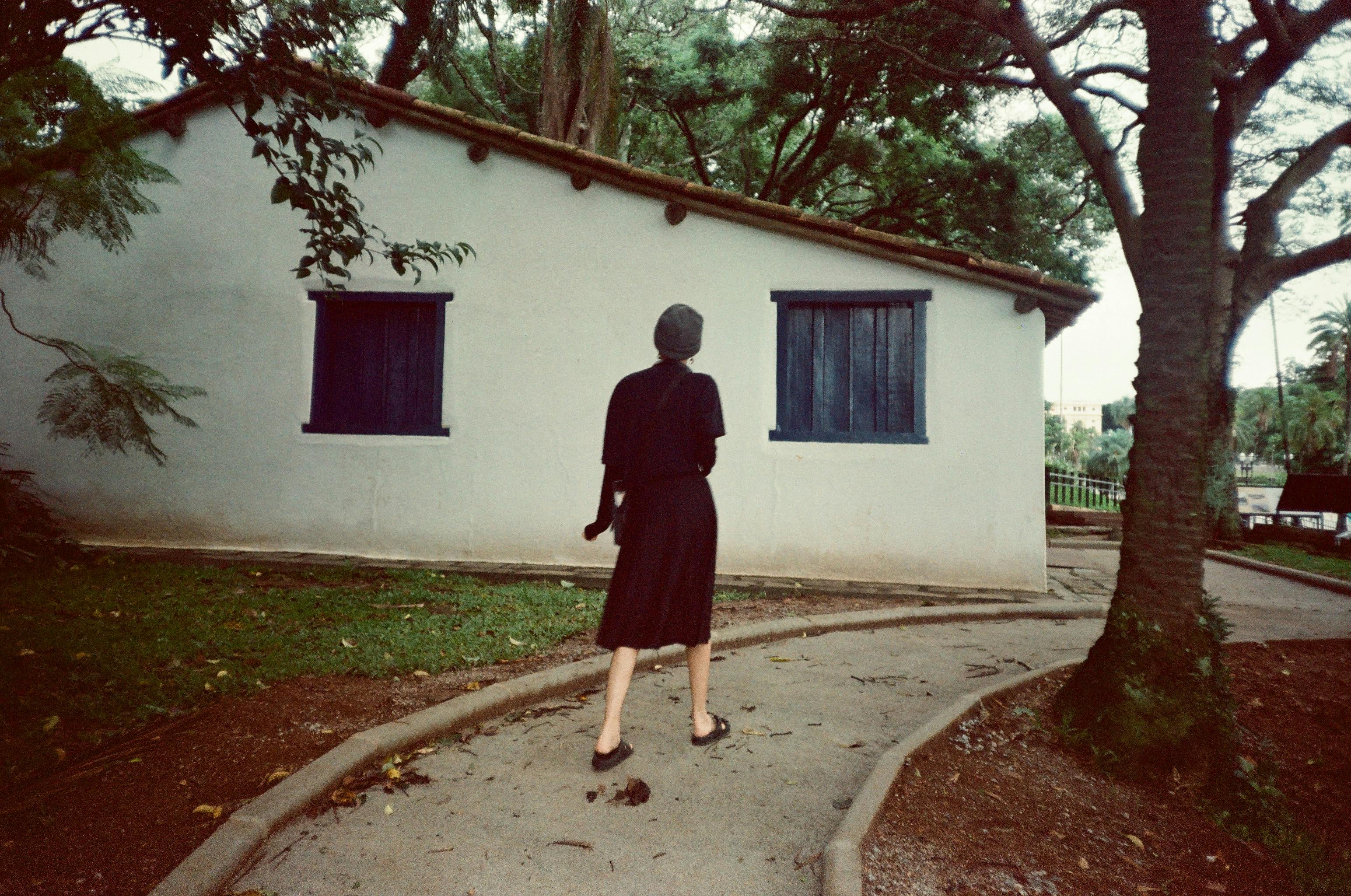 A woman in a black dress walks along a serene village path, surrounded by trees and a quaint house.