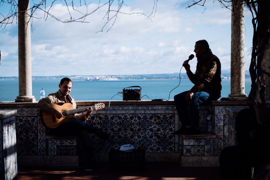 Two men perform music by the Lisbon seaside, one playing guitar and the other singing with a microphone.