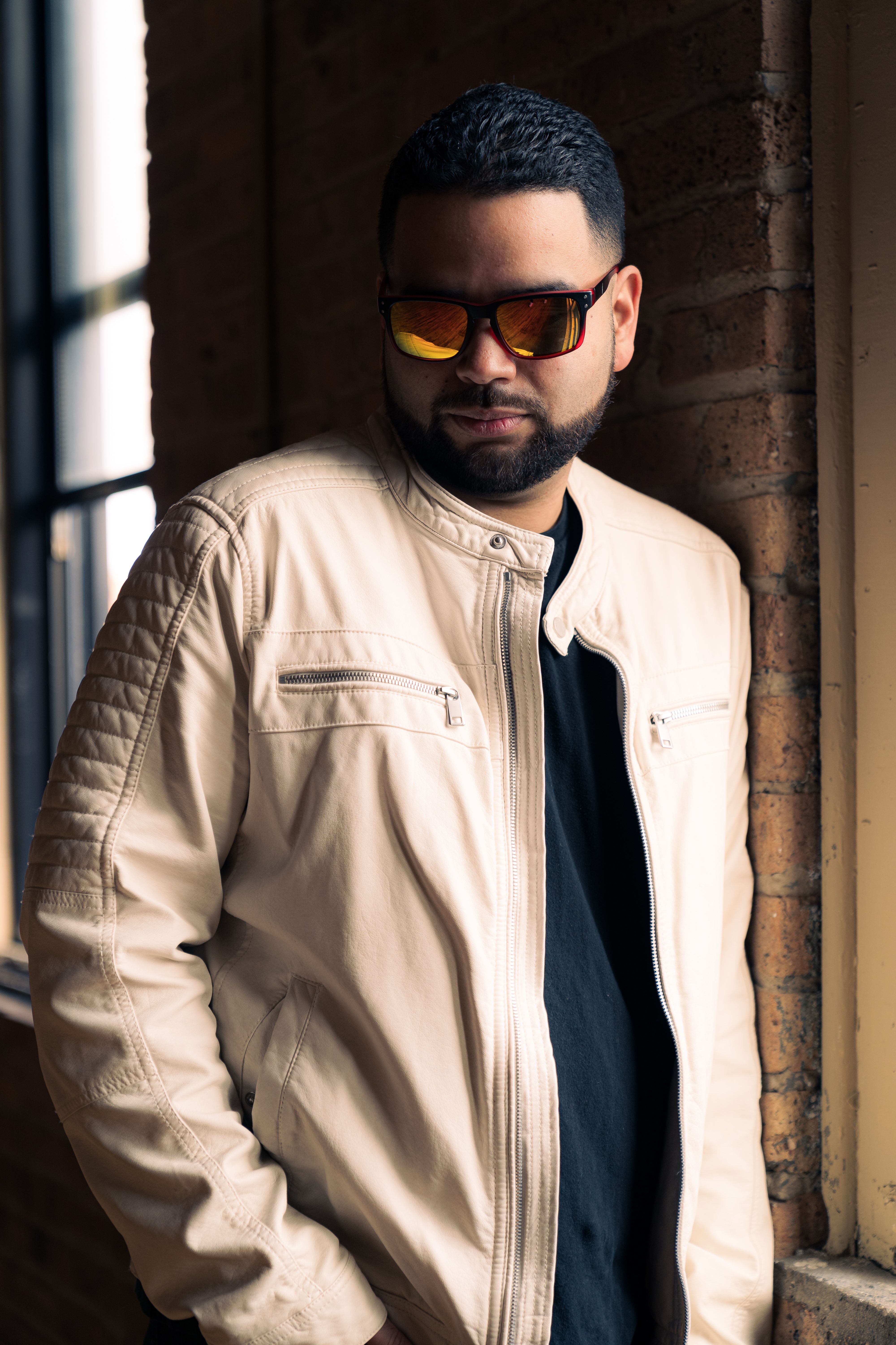 Stylish man with sunglasses and white leather jacket, leaning against an indoor brick wall.