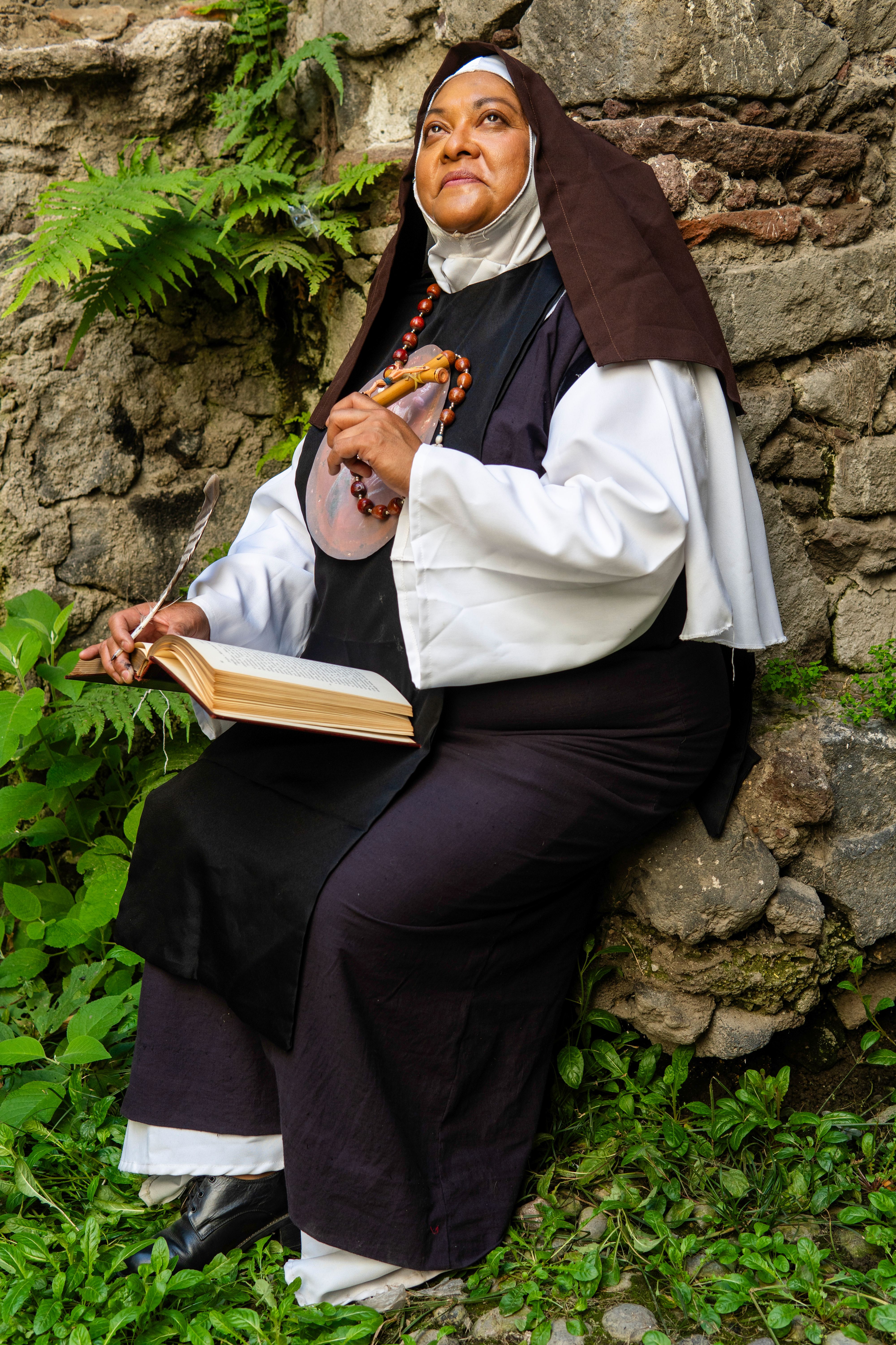 A contemplative nun with a rosary and bible, sitting by a stone wall in Mexico City.