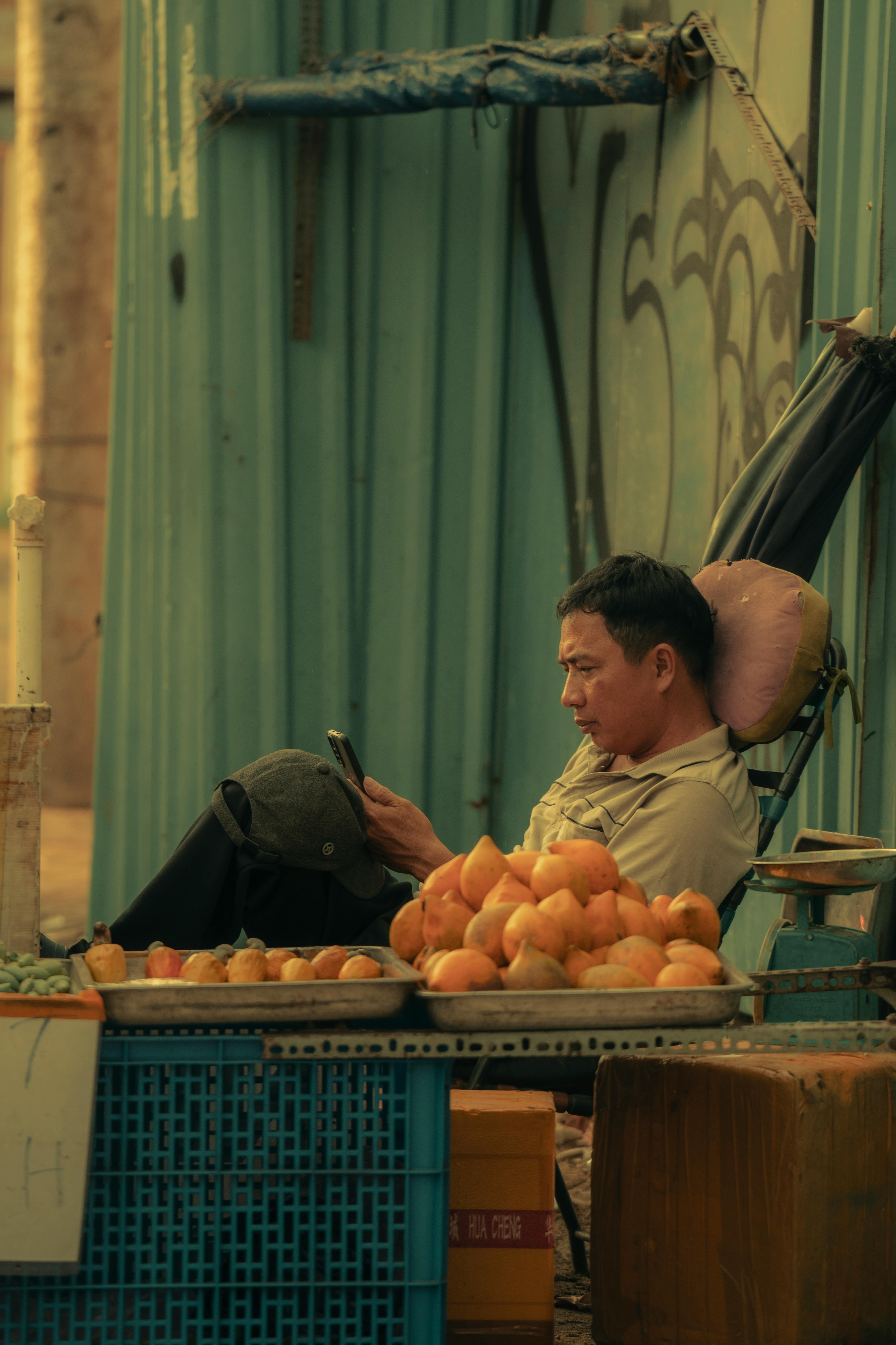 Asian man sitting at a fruit stand in a bazaar, using a smartphone. Urban outdoor scene.