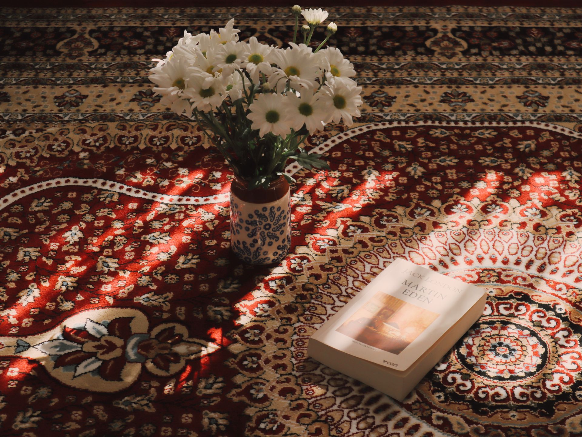 A cozy scene with white flowers in a vase and a book on a patterned carpet illuminated by sunlight.