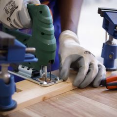 Gloved hands guiding a green jigsaw along a pine board held in blue clamps on a workbench