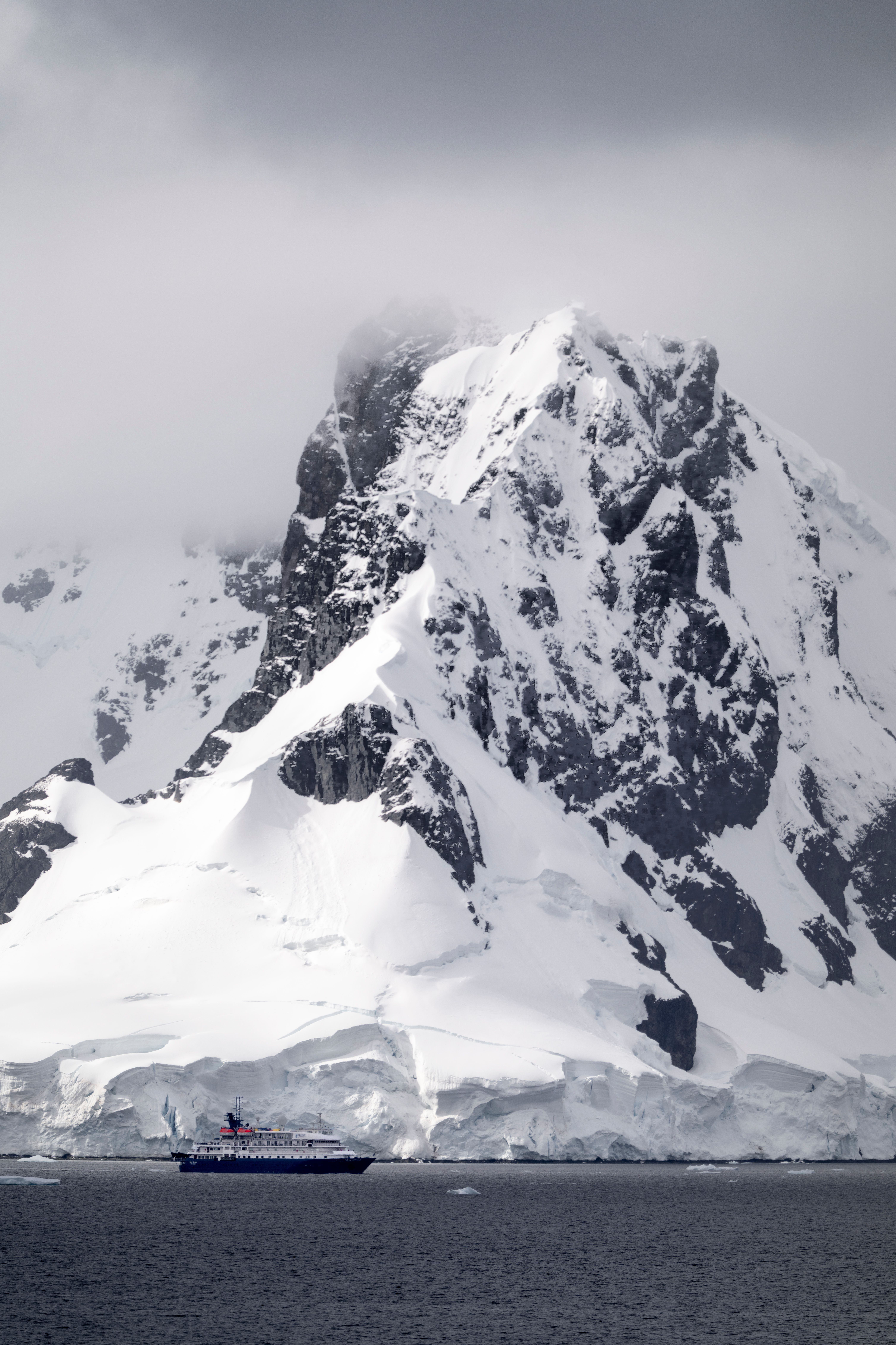 A ship navigates near a snowcapped mountain in Antarctica, under dramatic cloudy skies.