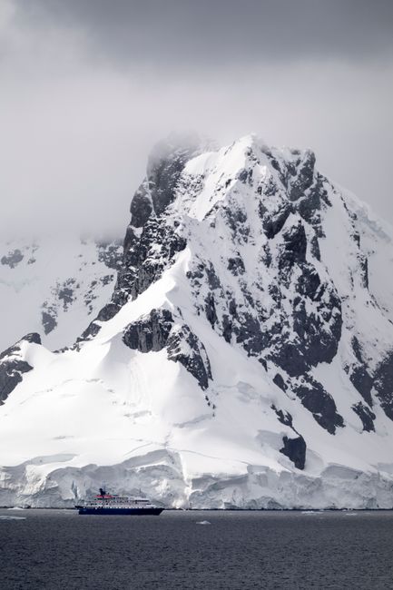 A ship navigates near a snowcapped mountain in Antarctica, under dramatic cloudy skies.