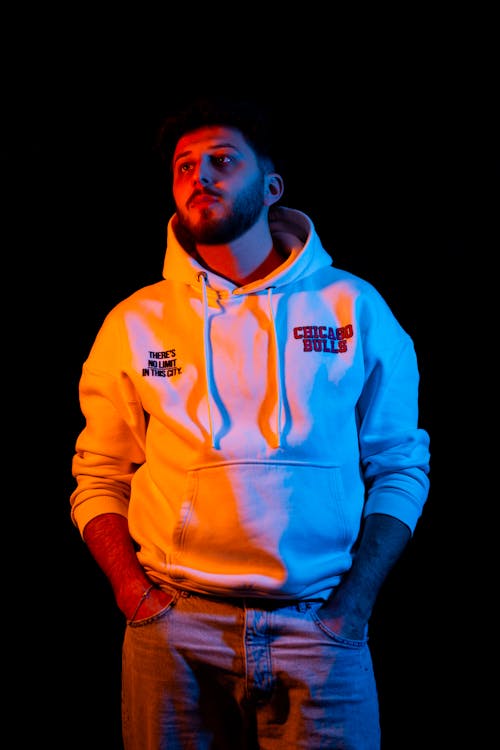 A man in a white hoodie and jeans standing in front of a black background