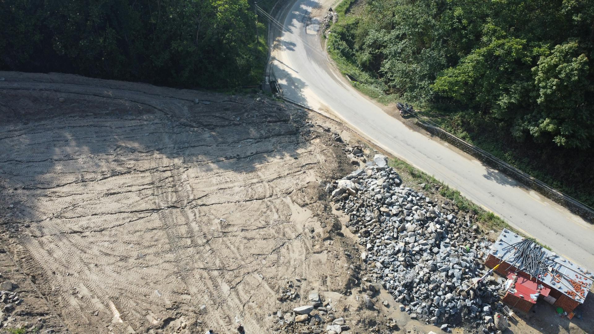 Aerial view showing a construction site with leveled ground, road, and surrounding forest in Panauti, Nepal.